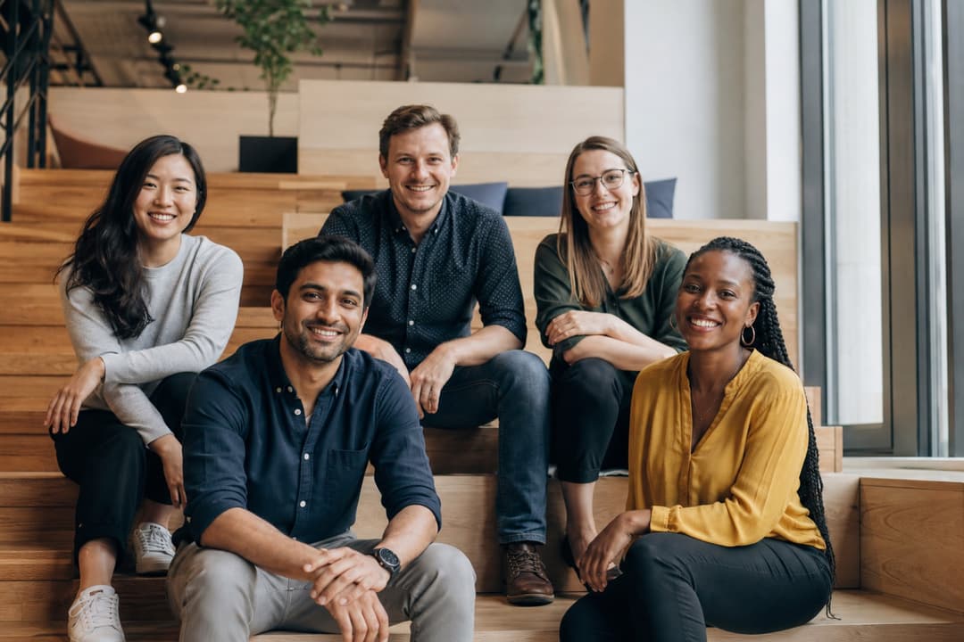 Five colleagues sitting on wooden office stairs, smiling toward the camera