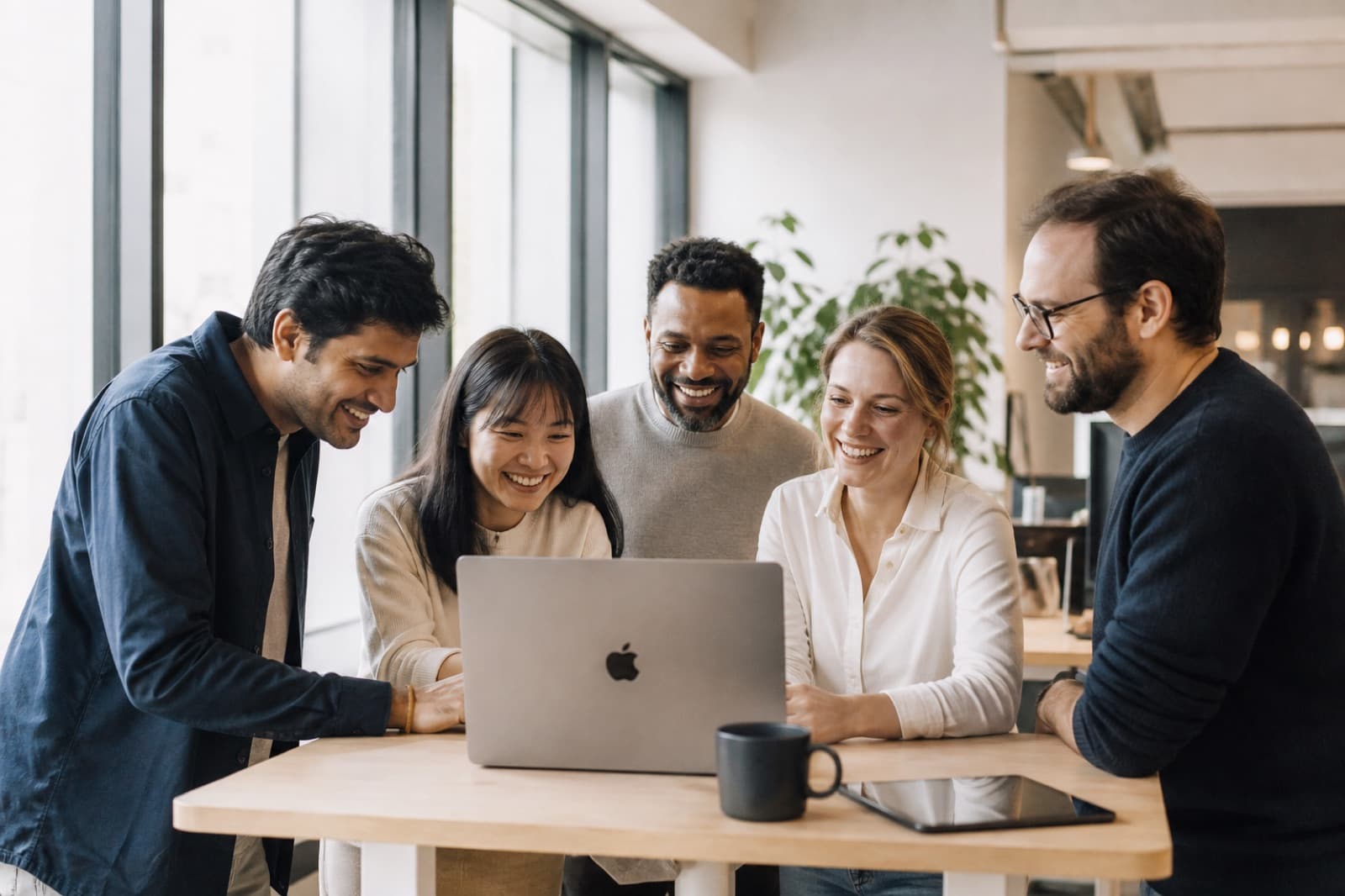 Five coworkers gathered around a standing desk, smiling at a laptop in a sunlit office